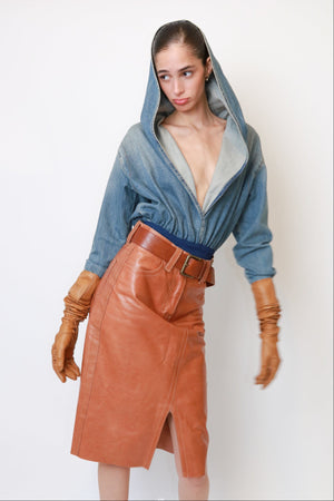 A model wearing a blue denim hooded top with a gray lining and a brown leather wrap skirt with a matching belt, styled with brown leather gloves against a plain white background.