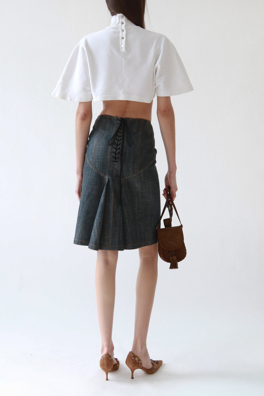 A model shown from the back wearing a white cropped short-sleeve blouse with a buttoned neck and a dark gray pleated denim skirt with a lace-up back. She is holding a brown leather handbag and wearing brown pointed-toe heels against a plain white studio background.