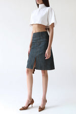 A model wearing a dark gray denim skirt with a front slit, paired with a white cropped short-sleeve top and brown pointed-toe heels, against a plain white studio background.