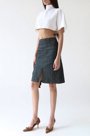 A model wearing a dark gray denim skirt with a front slit, paired with a white cropped short-sleeve top and brown pointed-toe heels, against a plain white studio background.