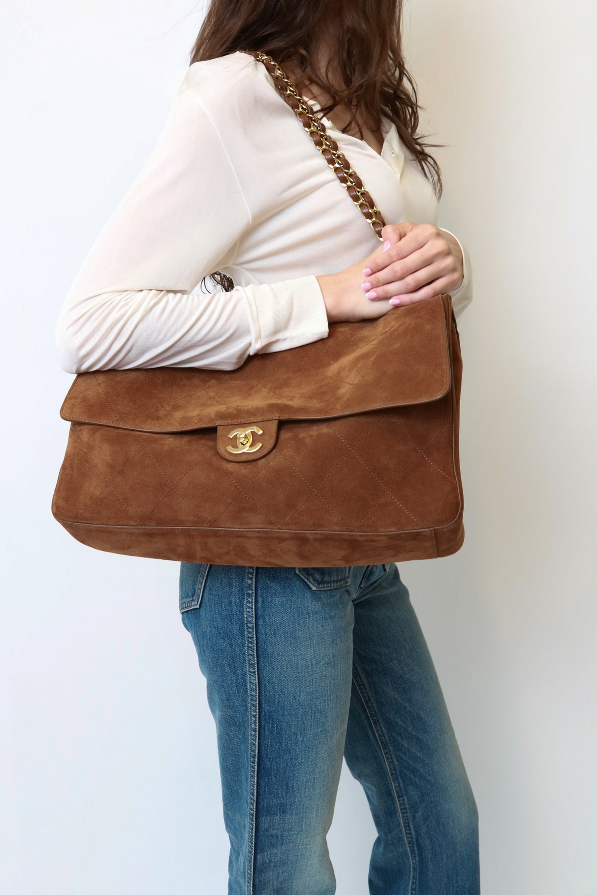 A woman wearing a white long-sleeve top and blue jeans carries a brown suede shoulder bag with a gold-tone interlocking CC logo clasp and a gold chain strap.