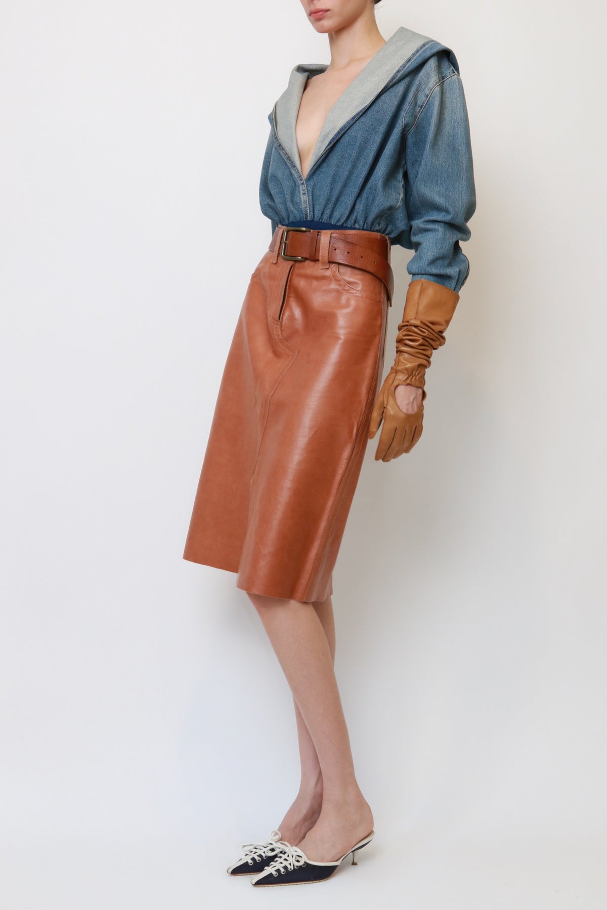A model is shown from the neck down wearing a brown leather midi skirt with a matching belt, a blue denim hooded top, brown leather gloves, and black pointed-toe mule heels with silver embellishments, set against a plain white studio background.