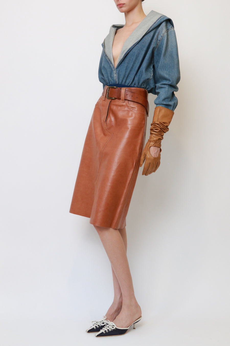A model is shown from the neck down wearing a brown leather midi skirt with a matching belt, a blue denim hooded top, brown leather gloves, and black pointed-toe mule heels with silver embellishments, set against a plain white studio background.