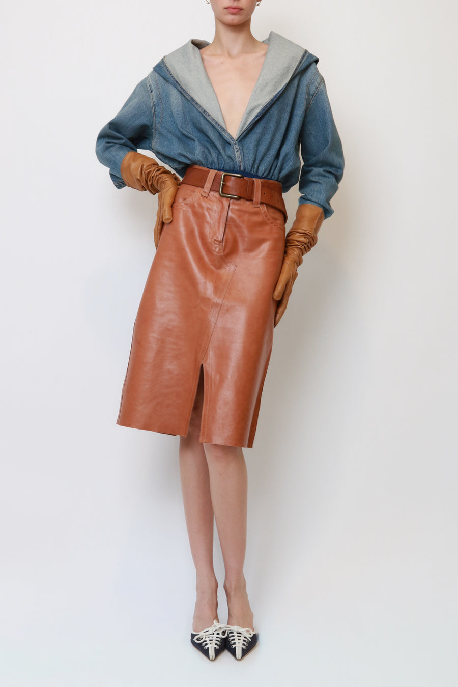 A model is shown from the neck down wearing a brown leather midi skirt with a front slit, a blue denim top with a gray hood, brown leather gloves, and black pointed-toe heels with white patterned detailing, set against a plain white studio background.