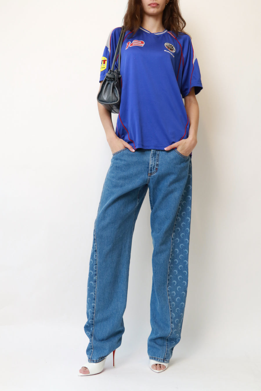 A model wearing a blue vintage short-sleeve sports jersey with '66' on the chest, paired with wide-leg blue jeans featuring a repeating wave pattern down the sides, white heeled sandals, and a black shoulder bag, set against a plain off-white background.